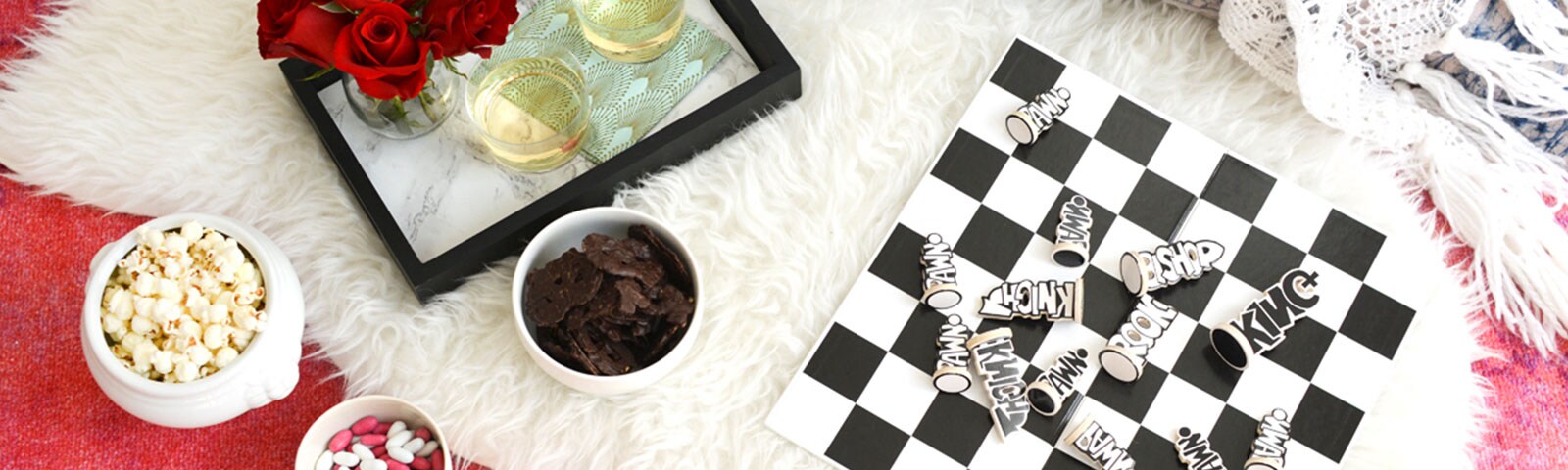 Atop a white shag rug sits a bowl of popcorn, a tray with roses and glasses of wine, a bowl of chocolate and a chess board. Atop a white shag rug sits a bowl of popcorn, a tray with roses and glasses of wine, a bowl of chocolate and a chess board.