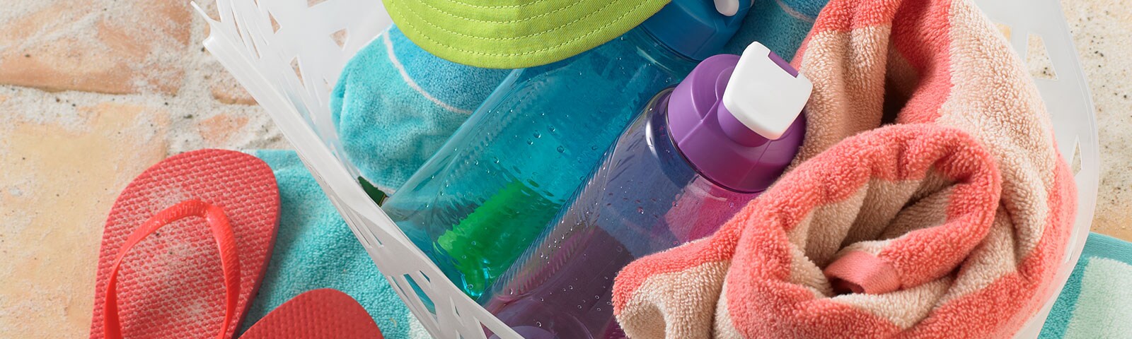 Close crop of a beach basket on sand packed with a towel, water bottles and a hat. To the left of the bag, flip flops.