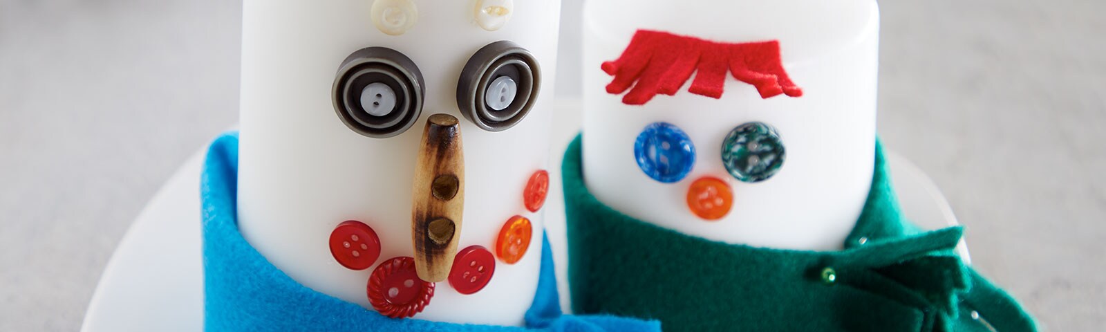 2 candles, each uniquely decorated with buttons and other craft items to look like snowmen wearing scarves