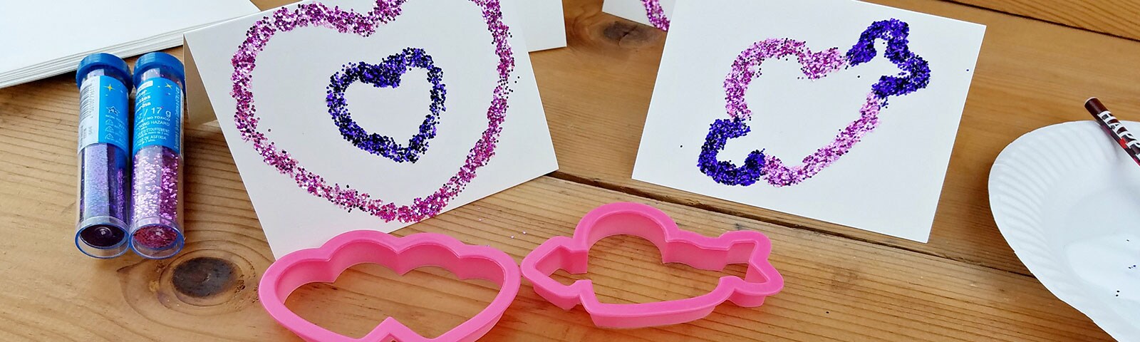 Wood table scattered with tubes of glitter, paper, and heart shaped cookie cutters as well as some completed Valentine's Day Cards