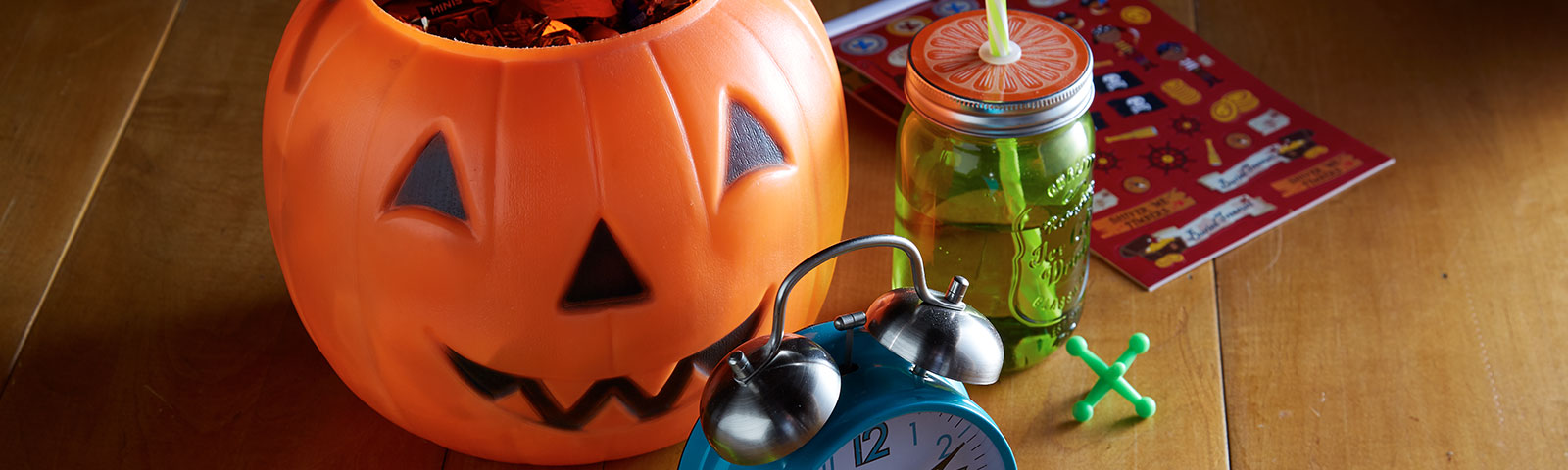 A jack-o-lantern candy bucket, a mason jar with lid and straw, and an alarm clock sitting on a wooden table